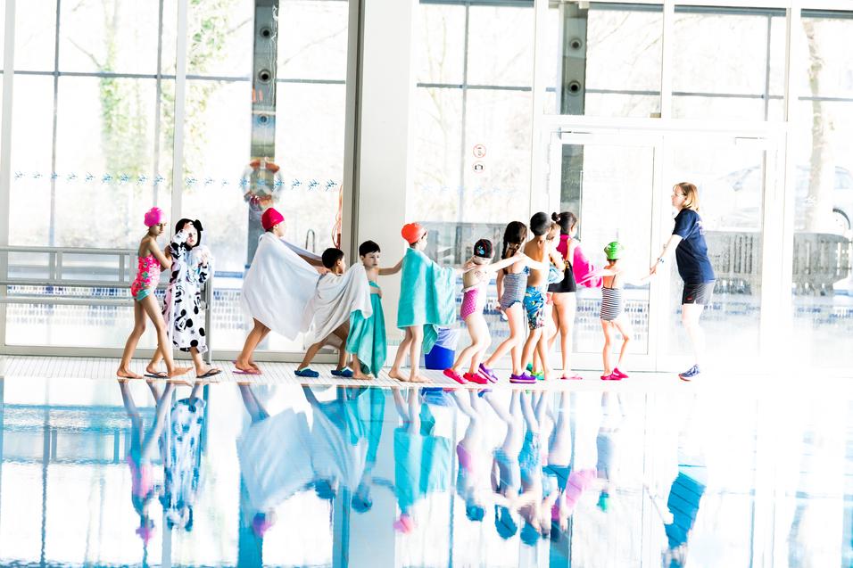 Kinder in Badesachen und Handtüchern bilden eine Schlange vor einem Hallenbad mit reflektierender Wasseroberfläche.